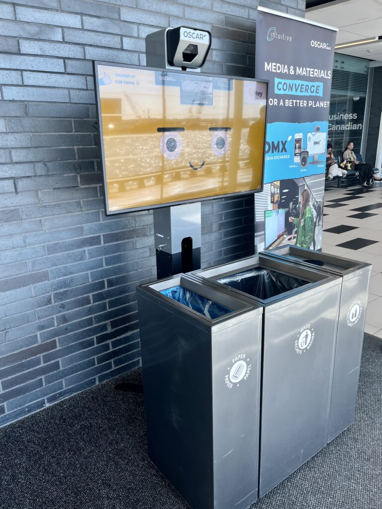Un écran Oscar Sort au-dessus de poubelles et de bacs de recyclage à l’aéroport.