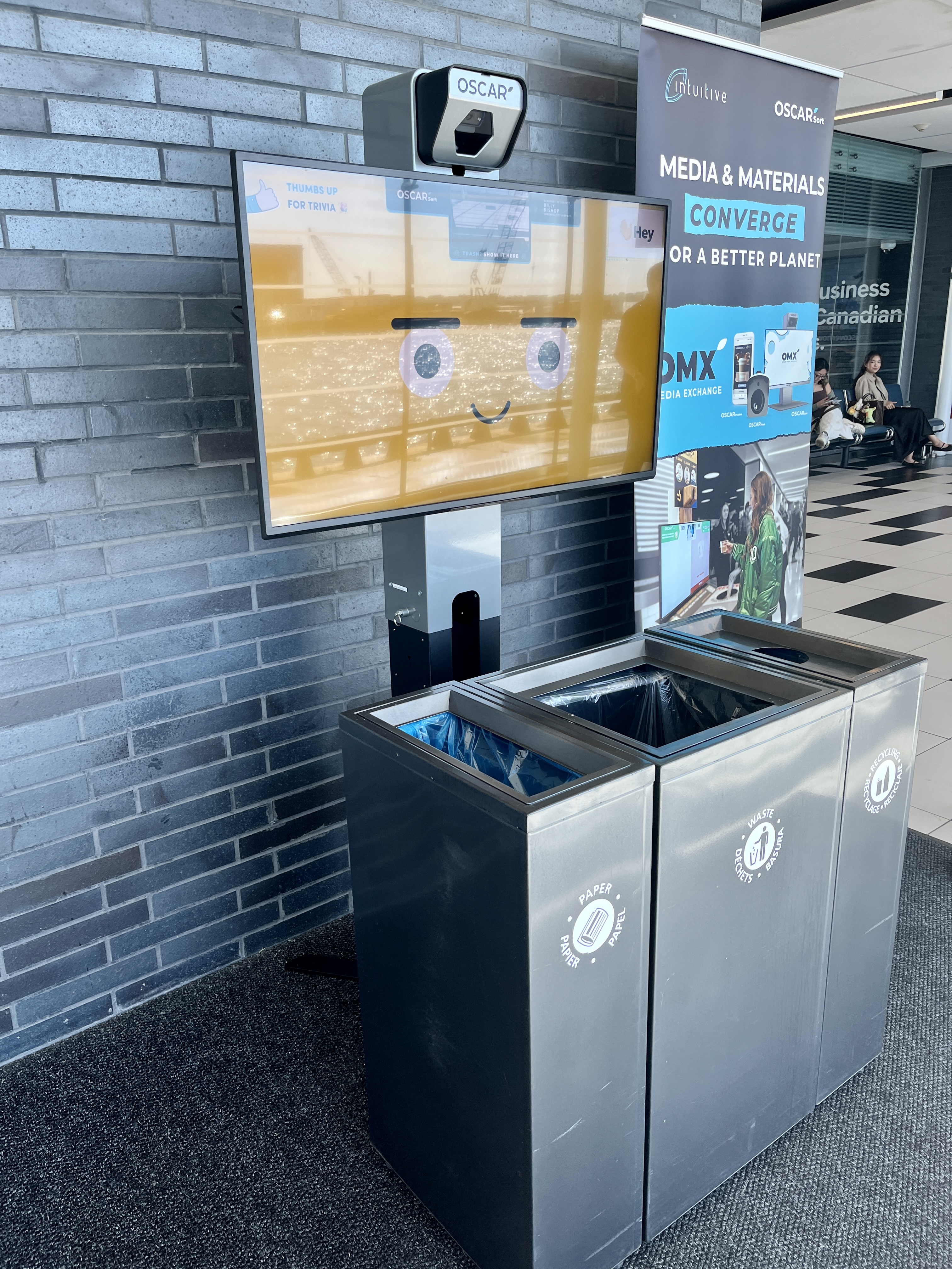 Un écran Oscar Sort au-dessus de poubelles et de bacs de recyclage à l’aéroport.